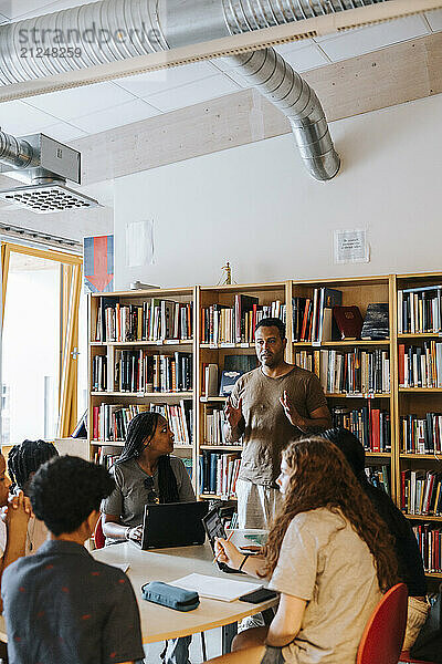 Male librarian explaining junior high students sitting near table in library at school