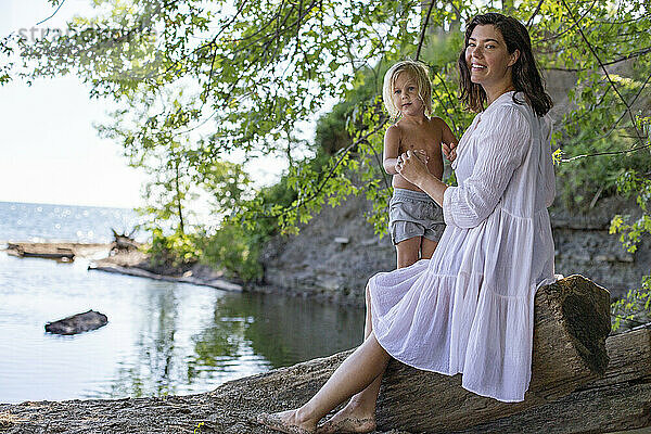 Woman in a white dress and her son sitting on a fallen tree by a lakeshore  surrounded by greenery on a sunny day.