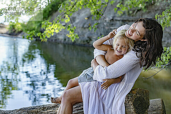 A joyful woman embraces a young boy by a serene lakeside  both smiling and enjoying a sunny day outdoors.