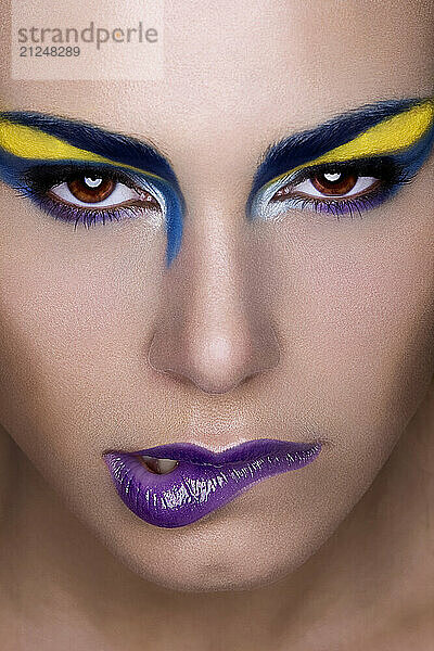 Close-up portrait of a person with bold yellow and blue eye makeup and glossy purple lipstick.