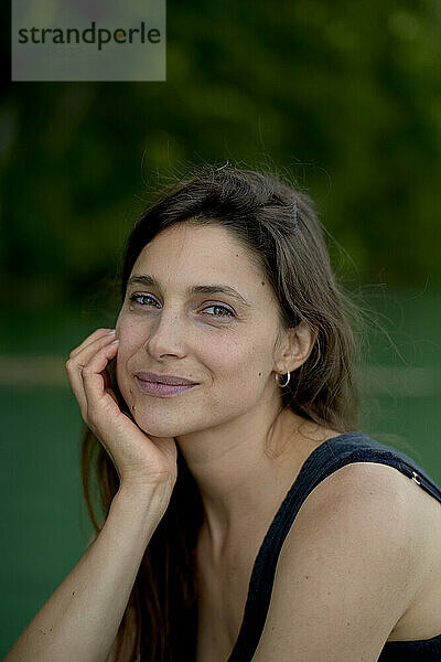 Portrait of a smiling woman outdoors with her hand on her cheek against a green background.