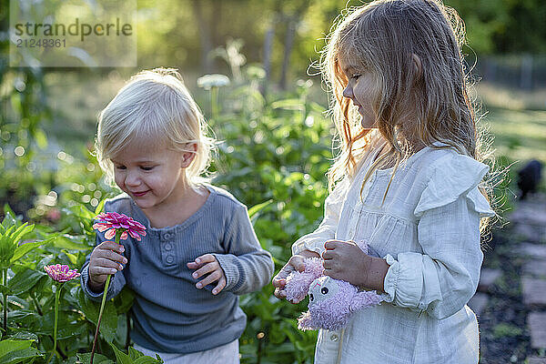 Two young girls enjoy the beauty and fragrance of a pink flower in a sunlit garden.