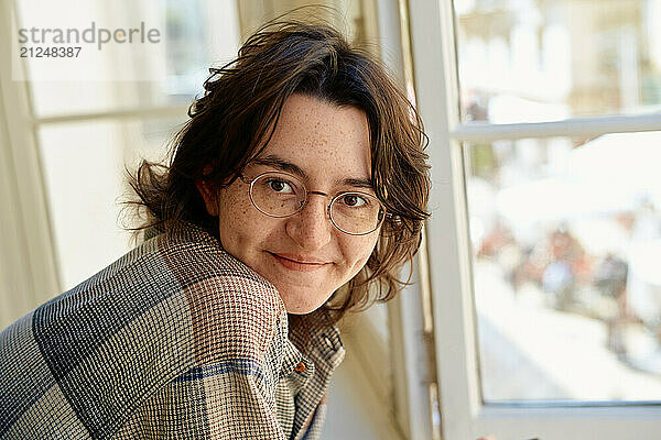 Smiling person with glasses leaning on a windowsill  looking at the camera with a street view in the background.