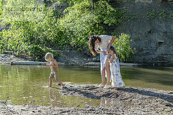 A woman and two children enjoy a sunny day by a lakeshore  with the woman bending down to interact with one of the children.