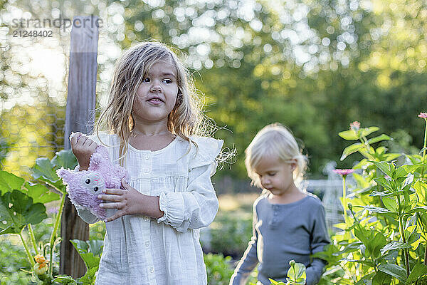 Young girl holding a pink stuffed animal stands in a garden while another girl walks behind her.