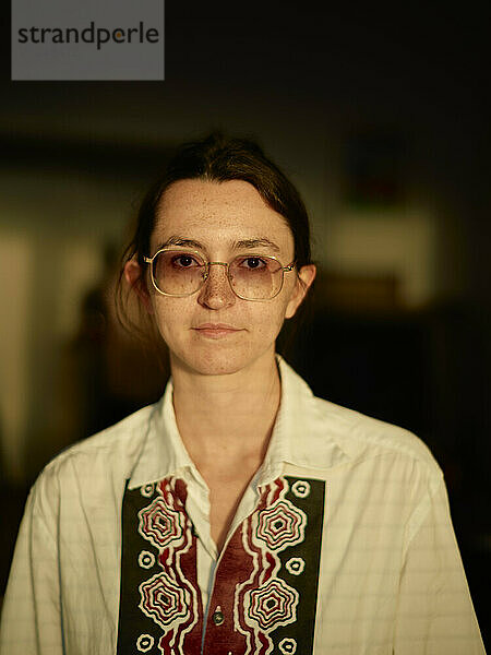 Portrait of a young woman wearing large round glasses and a white shirt with a red embroidered pattern standing against a blurred background.