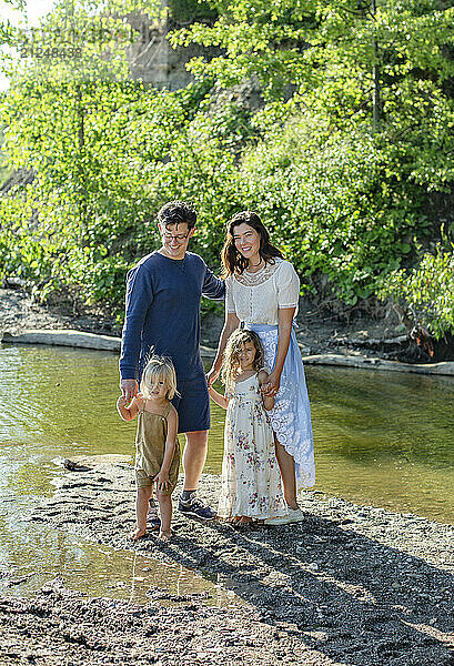 A family of four stands smiling on a sandy riverbank  with two adults and two young children enjoying a sunny day outdoors.