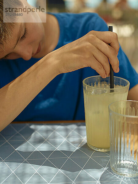 A teenager in a blue shirt stirring a glass of lemonade with a straw at a table.