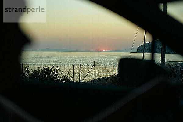 Sunset viewed through the silhouette of a car window overlooking the ocean and distant hills.