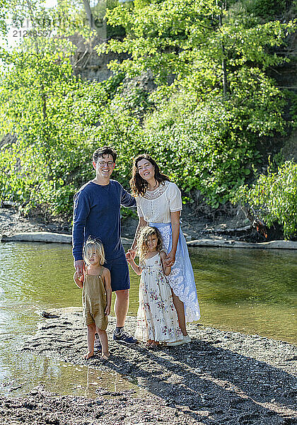 Family of four standing happily by a riverbank with a backdrop of trees and sandy terrain.