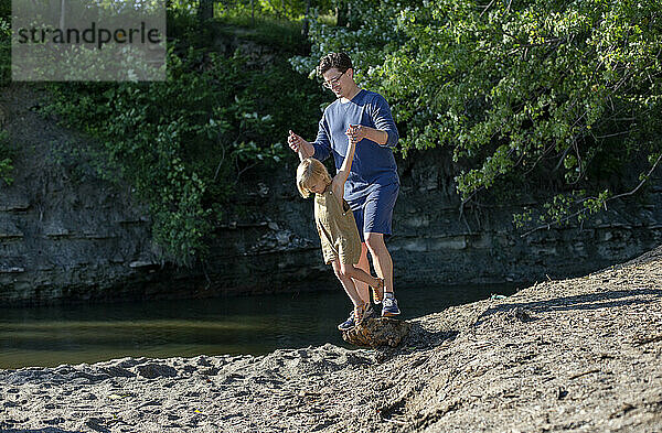 Adult male and child enjoying a playful moment on a sandy beach near a water body with greenery in the background.