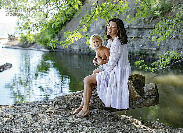 Woman in a white dress and her son sitting on a fallen tree by a lakeshore  surrounded by greenery on a sunny day.