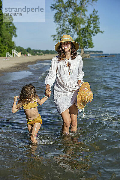 Woman and child holding hands and walking in the ocean shallows with smiles  the woman holding a sunhat in her free hand.