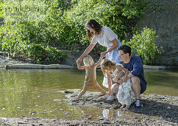 A family enjoys a sunny day playing by a riverside  with a young child taking tentative steps into the water under the watchful eyes of his parents.