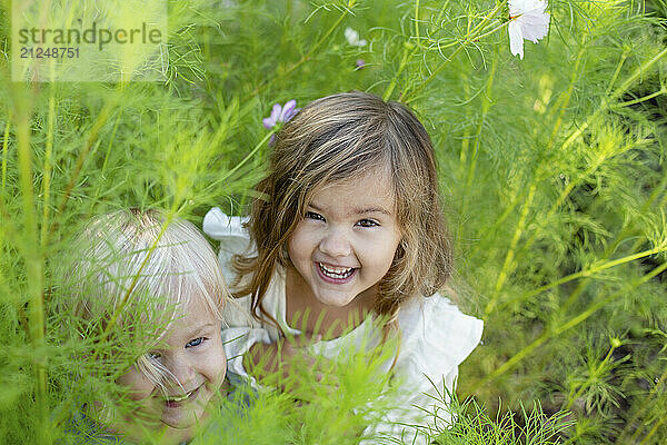 Two children smiling and peeking through lush green foliage with white flowers.