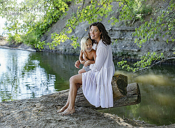 Woman in a white dress and her son sitting on a fallen tree by a lakeshore  surrounded by greenery on a sunny day.