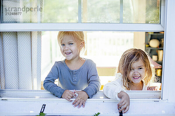 Two young girls smiling and looking through an open window on a sunny day.