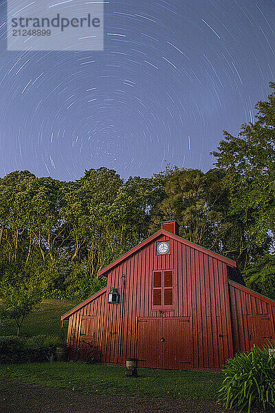 Star trails swirl above a charming red barn surrounded by trees at night  Auckland  New Zealand