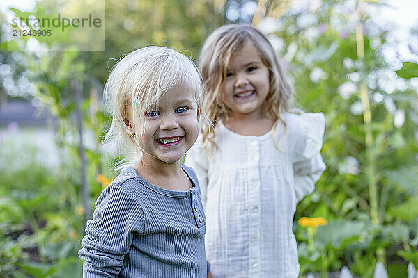 Two smiling young girls standing outdoors in a garden  with one girl embracing the other lightly.