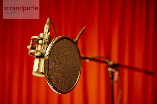 A professional studio microphone with a pop filter against a blurred red curtain background.