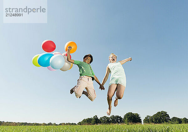 Children jumping with balloons on a sunny day holding hands.