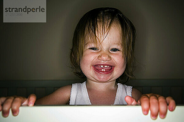 A 15-month-old girl is photographed smiling while standing in her crib