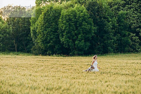 Young woman running through a rye field with a dog in summer