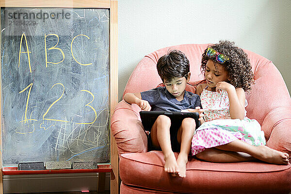 Two children sitting together on a pink chair  using a tablet  w