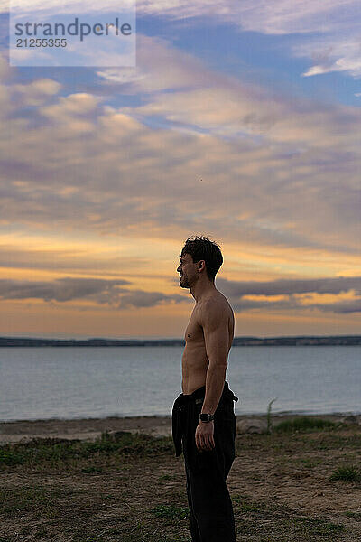 A man against the backdrop of the sunset sky on the shore of a lake.