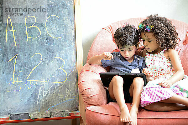 Two children sharing a tablet while sitting on a pink chair next