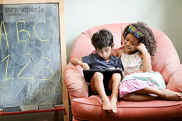 Two children sitting on a pink chair  using a tablet together  w