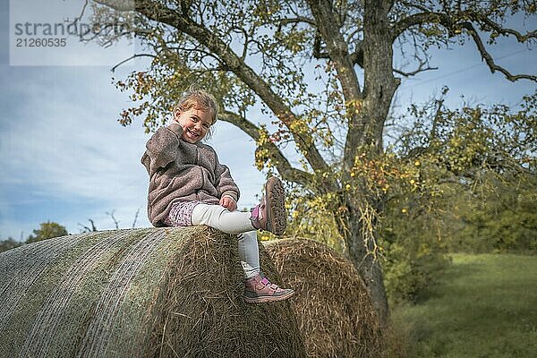 Kind sitzt fröhlich auf einem Strohballen in einer herbstlichen Landschaft mit Baum im Hintergrund