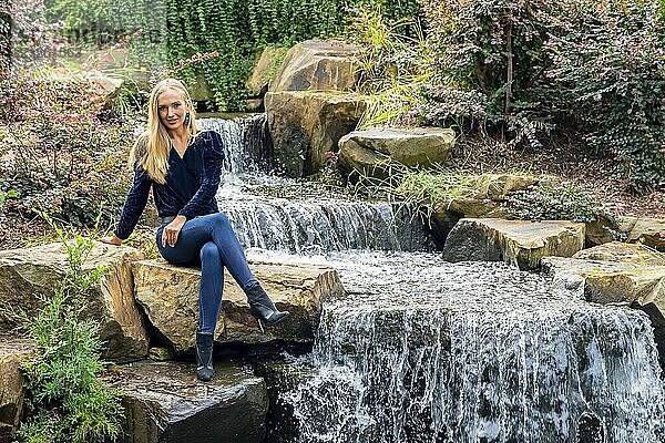 Blonde Schönheit glänzt in atemberaubender Herbstkleidung in der Nähe eines Wasserfalls im Park  eine malerische Herbstszene