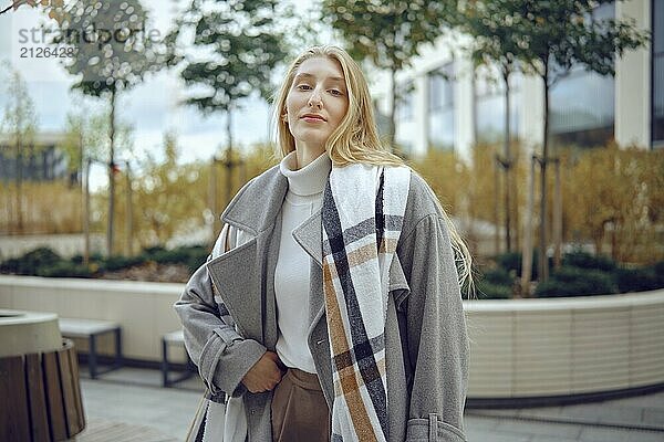 Eine junge Frau steht in einem modernen Stadtgarten und präsentiert ihr modisches Ensemble. Sie trägt einen gemütlichen Rollkragenpullover und einen übergroßen Mantel und strahlt Selbstbewusstsein inmitten von Grün und Stadtarchitektur aus