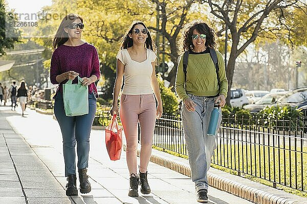 Eine Gruppe von drei Frauen  die an einem sonnigen Tag fröhlich im Freien spazieren gehen  Einkaufstaschen tragen und in Freizeitkleidung und mit Sonnenbrille gekleidet ihre Zeit miteinander genießen