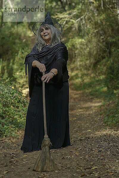 Alte schwarz gekleidete Hexe mit weißen Haaren auf einem Waldweg mit ihrem Besenstiel in der Hand