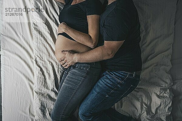 Ein Paar liegt im Bett  wobei der Mann die schwangere Frau umarmt und seine Hand auf den Babybauch legt