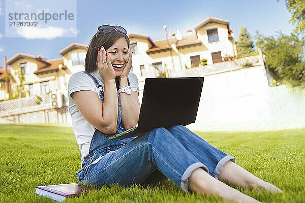 Frau bekommt freudige Emotionen durch Gespräche mit ihren Freunden auf einer Telefonkonferenz von Notebook  sie gekleidet in Jeans Overalls sitzen auf Gras im Park