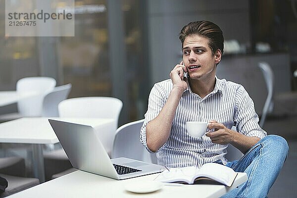 gutaussehender kaukasischer junger Mann mit Telefon  der am Laptop arbeitet und lächelt  während er einen Kaffee im Café genießt