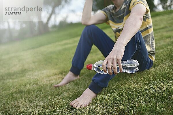 Junge kaukasische Mann Läufer entspannend hält Trinkwasserflasche und sitzt auf Gras im Park im Freien nach dem Sport am frühen Morgen Zeit  Bewegung und gesunde Konzept