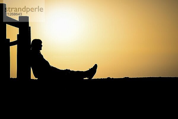 Silhouette einer unerkennbaren Person  die an einem Holzpfahl sitzt  mit orangefarbenem Sonnenuntergang im Hintergrund