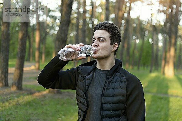 Ein gutaussehender junger Mann in einer Sportjacke  der in der Natur Wasser aus einer Flasche trinkt