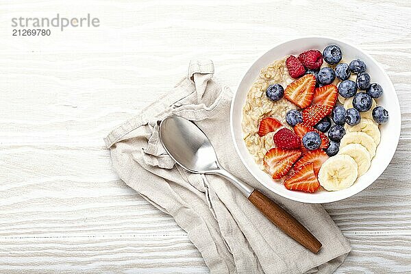 Haferflockenbrei mit Obst und Beeren in der Schüssel mit Löffel auf weißem Holzhintergrund Tischansicht  hausgemachte gesundes Frühstück Müsli mit Erdbeere  Banane  Heidelbeere  Himbeere. Platz für Text  Lebensmittel Fotografie  food photography