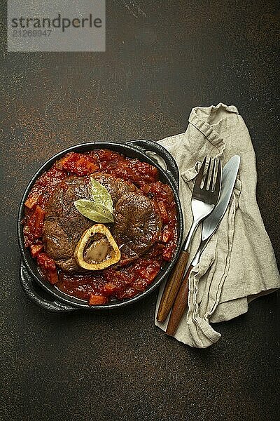 Traditionelles italienisches Gericht Ossobuco all Milanese aus geschnittener Kalbshaxe mit vegetarischer Tomatensoße  serviert in schwarzer Kasserolle von oben auf rustikalem braunem Hintergrund  Foodfotografie