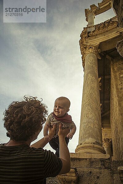 Mann mit lockigem braunem Haar hält ein Baby vor historischen Säulen eines alten römischen Tempels