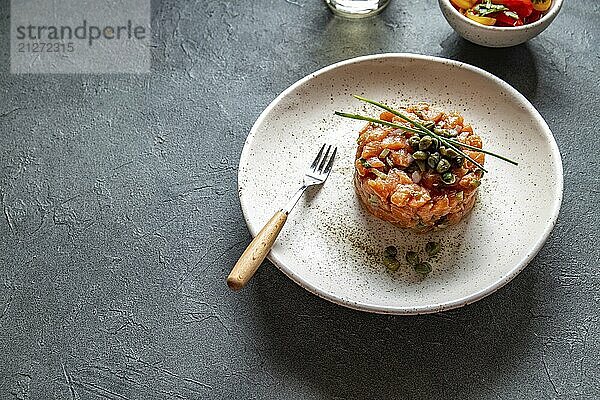 Lebensmittel  Lachs Tartar mit Kapern und lila Zwiebeln auf weißem Teller  grauer Hintergrund