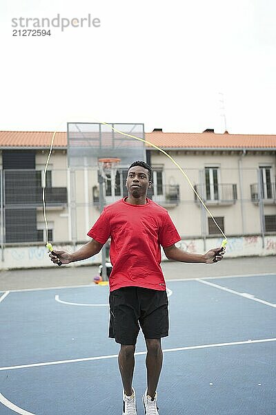 Vertikales Foto mit Kopierbereich eines jungen sportlichen afrikanischen jungen Mannes  der auf einem städtischen Basketballplatz im Freien Seil springt