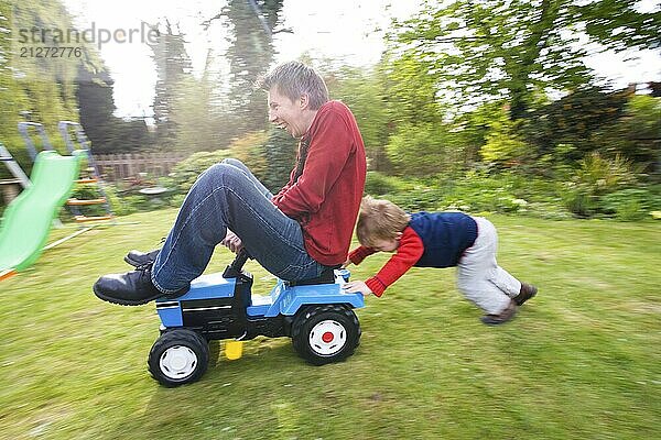 Der Sohn schiebt seinen Vater auf einem Spielzeugtraktor in ihrem Garten Themen der Erheiterung Familienzusammenhalt