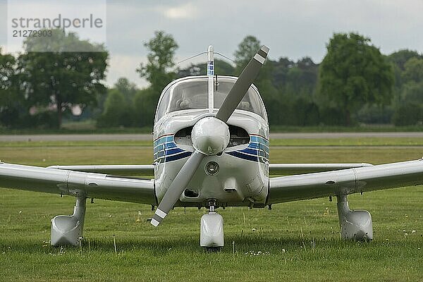 Vorderansicht eines einmotorigen weißen Propellerflugzeugs auf einer Wiese in der Nähe der Landebahn eines Flughafens