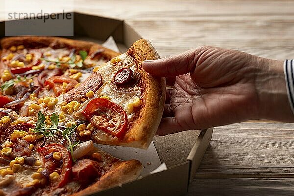 Menschliche kaukasische Hand nimmt ein Stück große Pizza aus einer Kartonschachtel auf einem weißen Holztisch